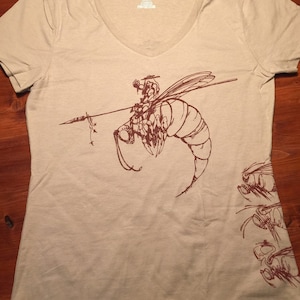 May include: A light beige t-shirt with a brown graphic of a bee with a rider on its back. The bee is flying and has a spear in its hand. The t-shirt has a v-neck and short sleeves.