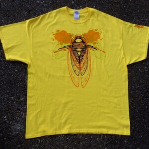 Pode incluir: Camiseta amarela com uma estampa gr&aacute;fica de um inseto cigarra com detalhes laranja e verde.