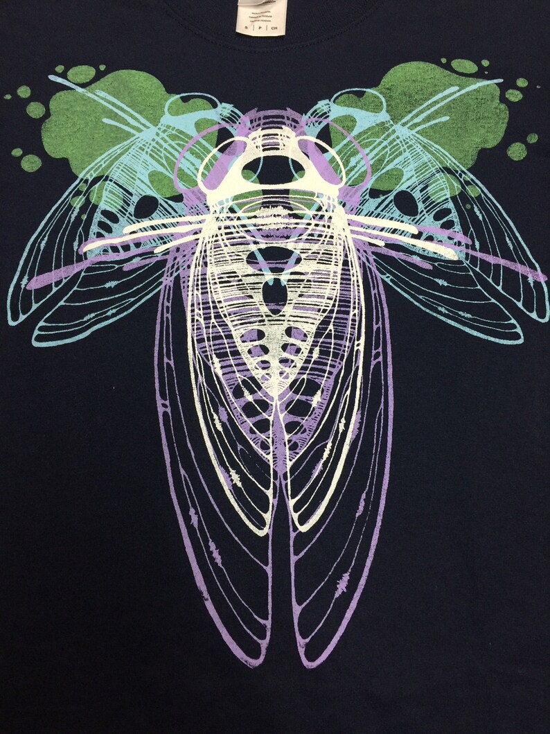 Pode incluir: Uma camiseta azul marinho com um gr&aacute;fico branco e roxo de uma cigarra com as asas abertas. A cigarra &eacute; delineada em branco e tem um corpo roxo. O fundo do gr&aacute;fico &eacute; de cor verde claro.