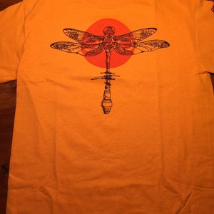 May include: Orange t-shirt with a black dragonfly design. The dragonfly is centered on the shirt and has a red circle behind it.
