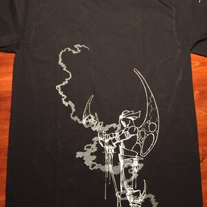 Op de afbeelding: Zwart T-shirt met een witte grafische print van een gestileerde figuur die een wapen vasthoudt en voor een rookwolk staat.