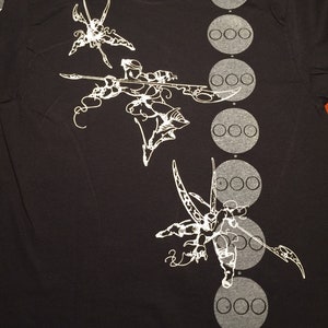 Op de afbeelding: Zwart T-shirt met een wit grafisch ontwerp van drie gestileerde figuren met lange ledematen en een rij van zes cirkels aan de rechterkant van het shirt.