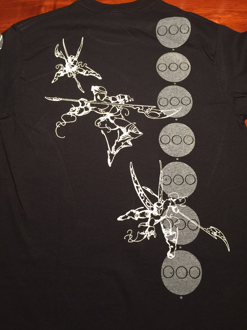 Op de afbeelding: Zwart T-shirt met een wit grafisch ontwerp van drie gestileerde figuren met lange ledematen en een rij van zes cirkels aan de rechterkant van het shirt.