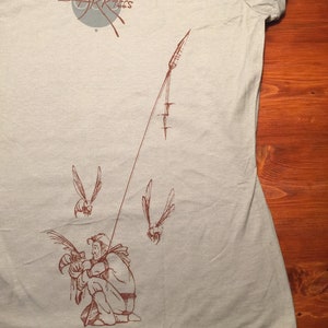 May include: A light blue t-shirt with a brown graphic of a person holding a bird. The graphic also includes a line with a spear at the end and two flying birds. The text "FULL ARK TIES" is printed in gray on the shirt.
