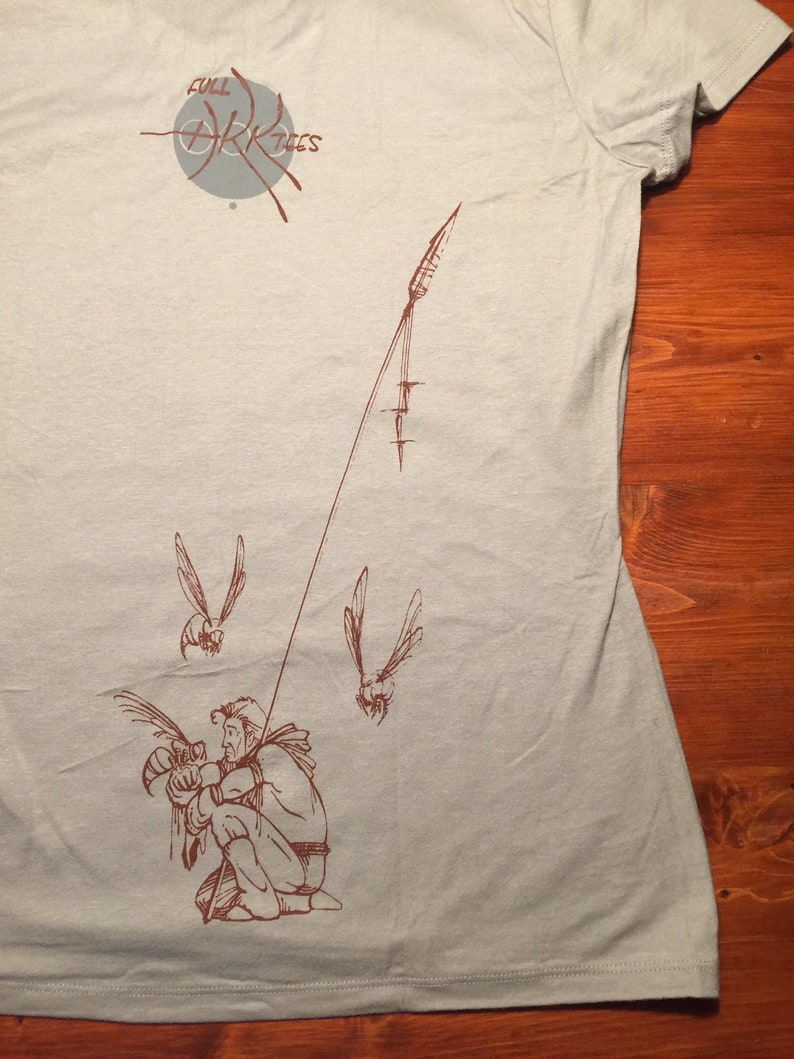May include: A light blue t-shirt with a brown graphic of a person holding a bird. The graphic also includes a line with a spear at the end and two flying birds. The text "FULL ARK TIES" is printed in gray on the shirt.