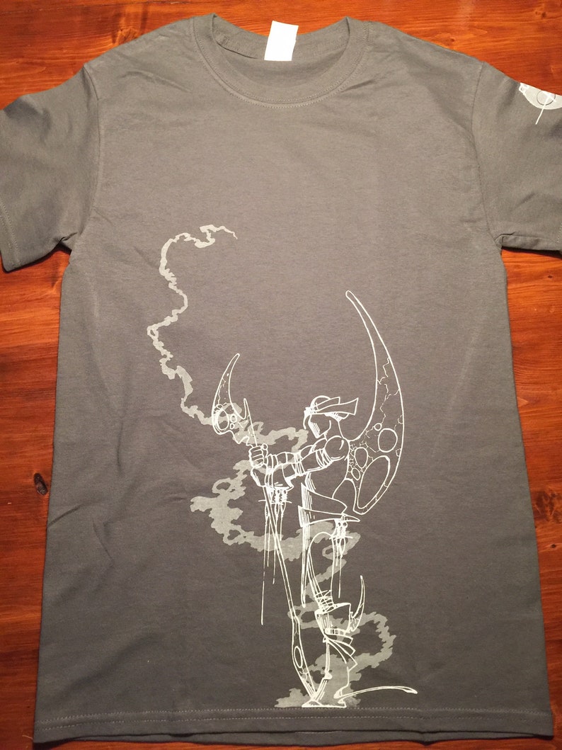 Op de afbeelding: Een grijs T-shirt met een witte afbeelding van een figuur die een halve maan vasthoudt en in een rookwolk staat.