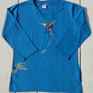 K&ouml;nnte beinhalten: Ein t&uuml;rkisfarbenes Langarmshirt mit einem goldenen Kolibri-Design. Der Kolibri fliegt mit ausgebreiteten Fl&uuml;geln und hat einen langen Schwanz. Auf dem linken &Auml;rmel befindet sich ein kleineres Kolibri-Design.