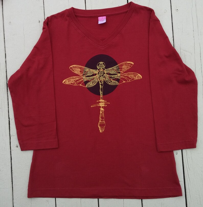 May include: A red long-sleeved shirt with a v-neck collar and a gold dragonfly graphic on the front.