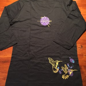 K&ouml;nnte beinhalten: Schwarzes Langarmshirt mit einem violetten Kreis und dem Text "Full Arktics" auf der R&uuml;ckseite. Die Vorderseite des Shirts hat ein gelbes Kolibri-Design mit violetten und blauen Akzenten.