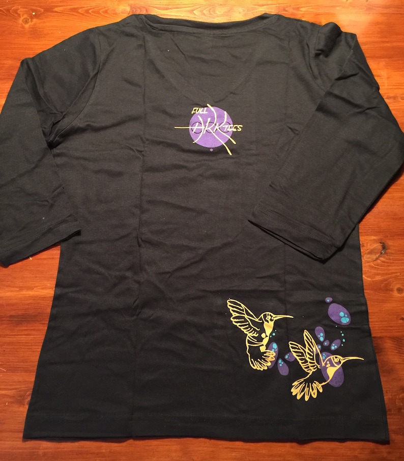 K&ouml;nnte beinhalten: Schwarzes Langarmshirt mit einem violetten Kreis und dem Text "Full Arktics" auf der R&uuml;ckseite. Die Vorderseite des Shirts hat ein gelbes Kolibri-Design mit violetten und blauen Akzenten.