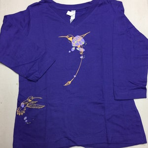 K&ouml;nnte beinhalten: Lilafarbenes Langarmshirt mit einem Kolibri-Motiv in Gelb und Orange. Der Kolibri fliegt mit ausgebreiteten Fl&uuml;geln und hat einen langen Schwanz. Auf der linken Seite des Shirts befindet sich ein kleineres Kolibri-Motiv.