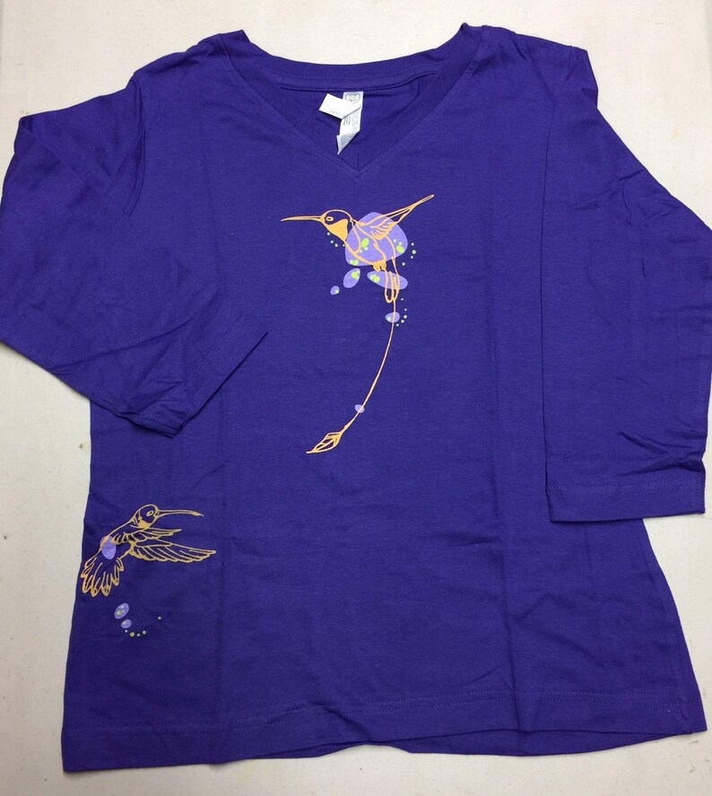 K&ouml;nnte beinhalten: Lilafarbenes Langarmshirt mit einem Kolibri-Motiv in Gelb und Orange. Der Kolibri fliegt mit ausgebreiteten Fl&uuml;geln und hat einen langen Schwanz. Auf der linken Seite des Shirts befindet sich ein kleineres Kolibri-Motiv.