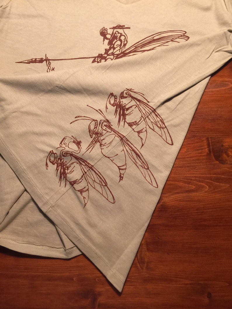 May include: A light green t-shirt with a brown graphic print of three insects and a figure holding a spear. The insects have large wings and detailed features.