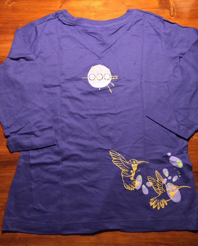 K&ouml;nnte beinhalten: Lilafarbenes Langarmshirt mit einem gelben Kolibri-Design und dem Text "Ooozes" auf der R&uuml;ckseite.