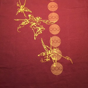 Op de afbeelding: Bordeauxrood T-shirt met een gele grafische print van een gestileerde krijgerfiguur met een zwaard en een reeks cirkels met drie stippen in elke cirkel.