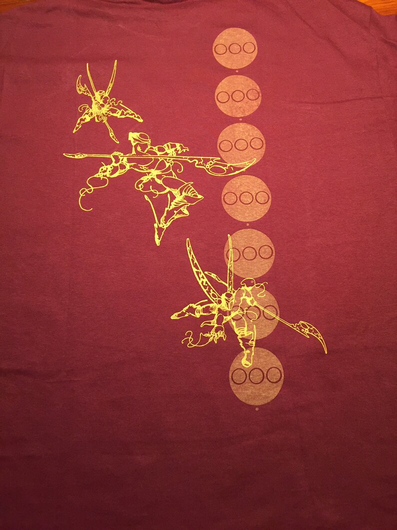 Op de afbeelding: Bordeauxrood T-shirt met een gele grafische print van een gestileerde krijgerfiguur met een zwaard en een reeks cirkels met drie stippen in elke cirkel.