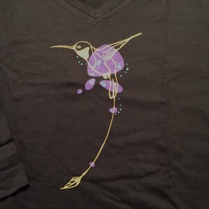 K&ouml;nnte beinhalten: Ein schwarzes T-Shirt mit einem gelben Kolibri-Motiv. Der Kolibri fliegt mit ausgebreiteten Fl&uuml;geln und hat lila und t&uuml;rkisfarbene Akzente.
