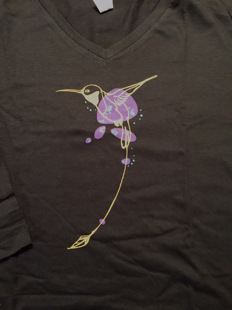 K&ouml;nnte beinhalten: Ein schwarzes T-Shirt mit einem gelben Kolibri-Motiv. Der Kolibri fliegt mit ausgebreiteten Fl&uuml;geln und hat lila und t&uuml;rkisfarbene Akzente.