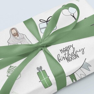 May include: Green and white wrapping paper with a green ribbon. The paper has a variety of illustrations including a person, a cupcake, a gift box, balloons, and a building. The text "Happy birthday Nixon" is written in black.