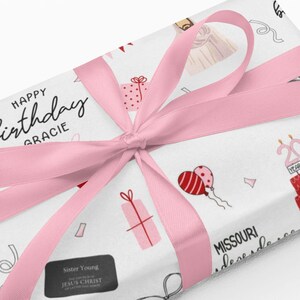 May include: Wrapped gift with a pink ribbon. The wrapping paper is white with birthday-themed illustrations, including a cake, balloons, and gift boxes. Text on the paper reads "Happy Birthday Gracie."
