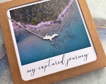 May include: A silver necklace with a pendant shaped like the Upper Peninsula of Michigan. The necklace is displayed in a brown box with a white card that says "my captured journey".