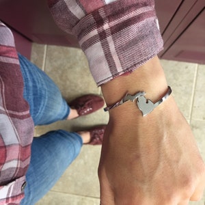 May include: A silver-colored cuff bracelet featuring a cutout design of the state of Michigan. The bracelet is worn on a wrist, with a plaid shirt and blue jeans visible in the background. The bracelet is a stylish accessory.