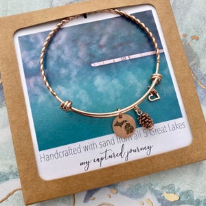 May include: A rose gold bracelet with a twisted band and a round charm featuring a state outline. The bracelet also has a heart charm and a pinecone charm. The card behind the bracelet says "Handcrafted with sand from all 5 Great Lakes" and "my captured journey".