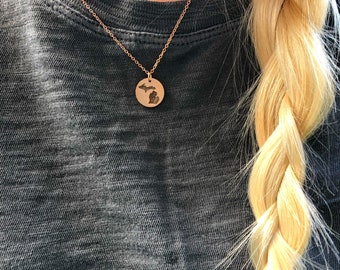 May include: A rose gold necklace with a round pendant featuring a detailed engraving of the state of Michigan. The pendant hangs from a delicate chain. The necklace is displayed against a gray background.