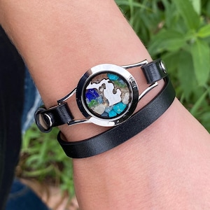May include: A black leather bracelet with a silver charm featuring a state outline filled with colorful sand and stones.