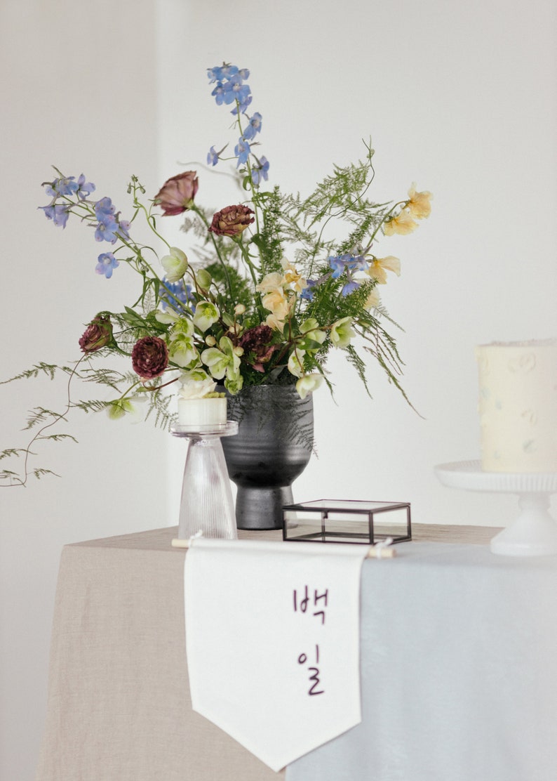 Puede incluir: Un arreglo floral con flores azules, amarillas y burdeos en un jarr&oacute;n oscuro. Un pastel peque&ntilde;o est&aacute; sobre un soporte de vidrio transparente. Una pancarta con texto coreano est&aacute; sobre la mesa.