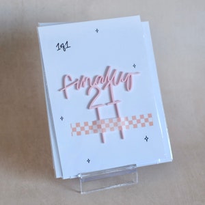 May include: Pink acrylic cake topper with the text "Finally 21" and a chequered ribbon. The topper is on a white card with the text "1 of 1" written on it.