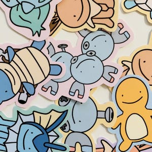 Op de afbeelding: Een verzameling kleurrijke cartoon karakter stickers in verschillende vormen en maten. De stickers tonen wezens in tinten blauw, geel, oranje en groen, met zwarte contouren. De stickers zijn in een willekeurig patroon gerangschikt.