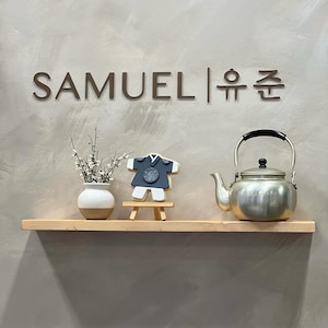 May include: A wooden shelf with a silver teapot, a small white vase with white flowers, and a black and white cookie shaped like a traditional Korean hanbok. The shelf is mounted on a light gray wall with the text "SAMUEL|유준" above it.