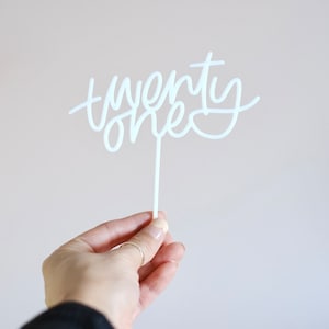 May include: White acrylic cake topper with the words "twenty one" written in a cursive script.