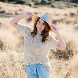 May include: A woman wearing a light brown crochet top with a wide-brimmed straw hat with a blue ribbon, blue denim shorts, and a white belt. She is standing in a field of tall grass.