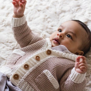 May include: A baby wearing a light brown and white knitted cardigan with wooden buttons. The cardigan has a pocket on the front. The baby is lying on a white fluffy surface.