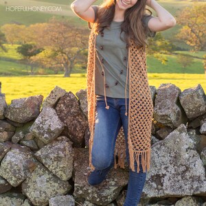 Peut inclure: Une femme portant un long gilet en crochet de couleur marron clair avec des franges. Elle se tient devant un mur en pierre avec un champ vert et des collines en arri&egrave;re-plan. Le gilet est orn&eacute; d'un collier de perles brun.