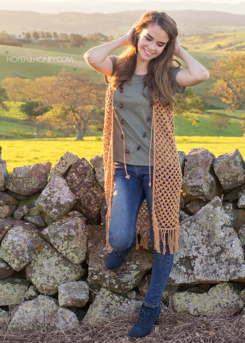 Peut inclure: Une femme portant un long gilet en crochet de couleur marron clair avec des franges. Elle se tient devant un mur en pierre avec un champ vert et des collines en arri&egrave;re-plan. Le gilet est orn&eacute; d'un collier de perles brun.