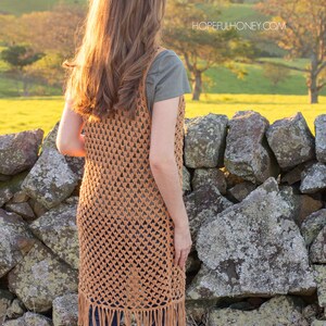 Peut inclure: Un long gilet crochet&eacute; avec une frange sur le bas, de couleur brun clair. Le gilet est port&eacute; sur un t-shirt vert et un jean bleu. La personne est debout devant un mur en pierre avec un champ vert et des arbres en arri&egrave;re-plan. Le texte "HOPEFULHONEY.COM" est visible dans le coin sup&eacute;rieur droit de l'image.