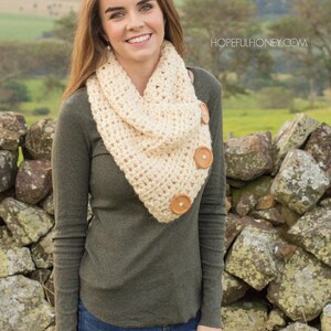 May include: A woman wearing a beige crocheted infinity scarf with wooden buttons. The scarf is worn over a green long-sleeved shirt and blue jeans. The woman is standing in front of a stone wall.