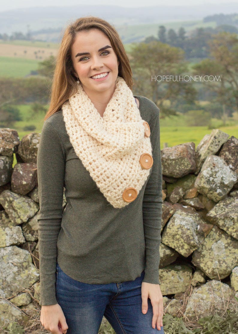 May include: A woman wearing a beige crocheted infinity scarf with wooden buttons. The scarf is worn over a green long-sleeved shirt and blue jeans. The woman is standing in front of a stone wall.