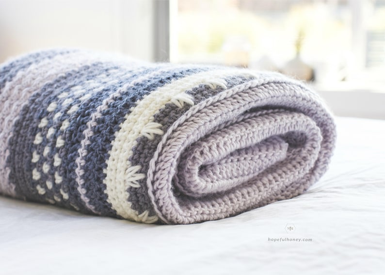 Pode incluir: Um cobertor macio de croch&ecirc; em tons de roxo, cinza e branco. O cobertor est&aacute; enrolado e colocado sobre uma superf&iacute;cie branca.