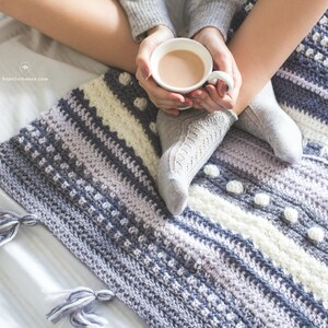 Pode incluir: Uma pessoa est&aacute; sentada em uma cama branca com um cobertor de croch&ecirc; em tons de cinza, branco e roxo. O cobertor tem um padr&atilde;o texturizado e pompons. A pessoa est&aacute; segurando uma caneca branca com uma bebida quente.