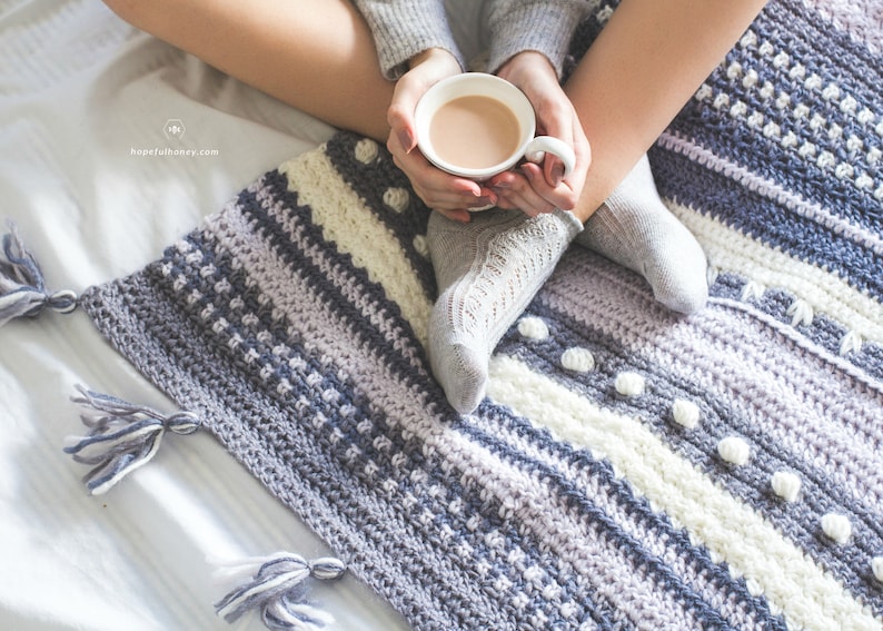 Pode incluir: Uma pessoa est&aacute; sentada em uma cama branca com um cobertor de croch&ecirc; em tons de cinza, branco e roxo. O cobertor tem um padr&atilde;o texturizado e pompons. A pessoa est&aacute; segurando uma caneca branca com uma bebida quente.