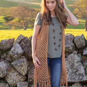 Peut inclure: Un long gilet crochet&eacute; avec des franges de couleur brun clair. Le gilet est port&eacute; sur un t-shirt vert et un jean bleu. Le mod&egrave;le est debout devant un mur en pierre avec un champ vert et des arbres en arri&egrave;re-plan.