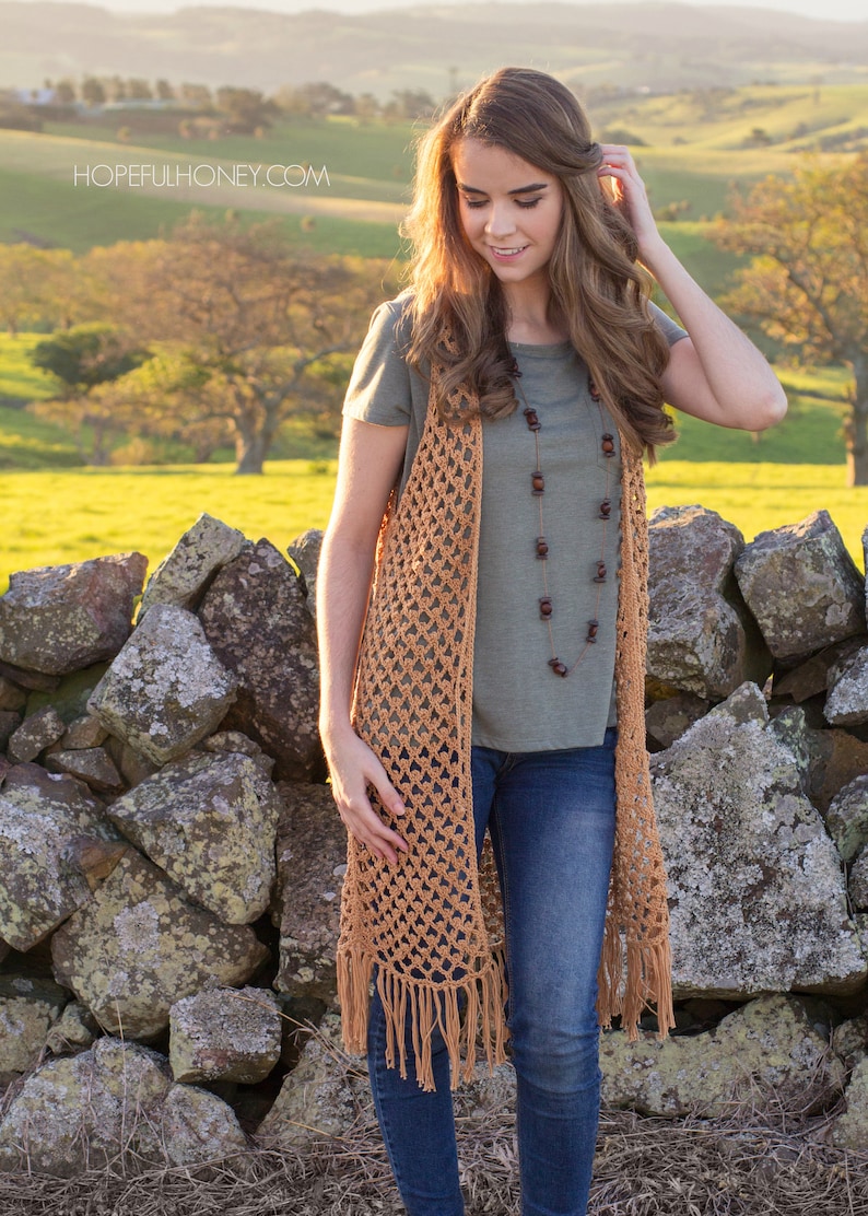 Peut inclure: Un long gilet crochet&eacute; avec des franges de couleur brun clair. Le gilet est port&eacute; sur un t-shirt vert et un jean bleu. Le mod&egrave;le est debout devant un mur en pierre avec un champ vert et des arbres en arri&egrave;re-plan.