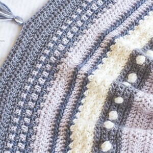 Pode incluir: Uma manta de croch&ecirc; com listras de fio cinza, roxo e branco. A manta tem um padr&atilde;o texturizado e pompons de fio branco.
