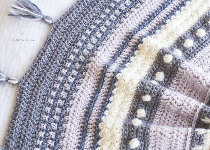 Pode incluir: Uma manta de croch&ecirc; com listras de fio cinza, roxo e branco. A manta tem um padr&atilde;o texturizado e pompons de fio branco.