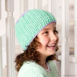 Puede incluir: Una persona joven lleva una gorra de punto de color verde claro con un borde morado y blanco. La gorra está hecha con un punto texturizado y tiene un diseño sencillo.