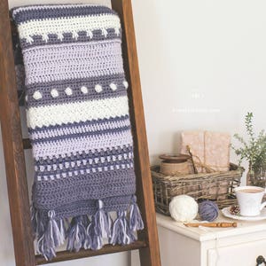 Pode incluir: Uma manta de croch&ecirc; com um padr&atilde;o listrado em tons de roxo e cinza, com detalhes em branco. A manta tem uma borda de franjas e est&aacute; pendurada em uma escada de madeira. A manta &eacute; feita &agrave; m&atilde;o e tem um ambiente aconchegante e r&uacute;stico.